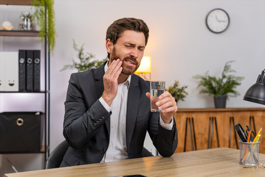 Middle-aged businessman at home office drinks cold water feels sharp tooth pain and searches dentist on smartphone. Freelancer guy at table presses cheek sighs hoping to book urgent clinic visit soon.
