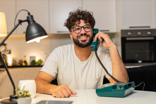 Arabian man at home on hotline talks on corded telephone listens asks brief questions solves issue. Egyptian guy sitting at table writes ticket on laptop gives steps polite voice calm support tonight.
