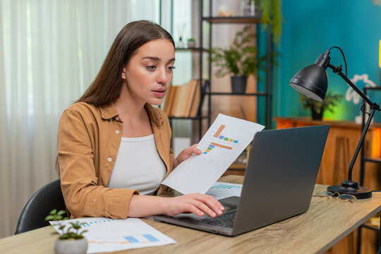 Young woman sitting at home table types notes on laptop while reviewing documents charts and complex data with serious focus. Caucasian girl interprets graphs and records findings for detailed report.
