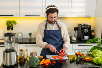 Middle-aged man chops tomatoes for fresh salad while wearing headphones singing in home kitchen. Young guy feels happy follows healthy diet for weight loss meal prep plus simple cooking for fitness. © Andrii Iemelianenko