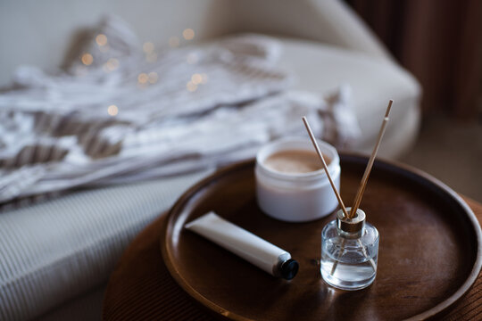 Home aroma liquid glass bottle diffuser with sticks on wooden tray and hand lotion in living room over couch. Hygge cozy atmosphere.