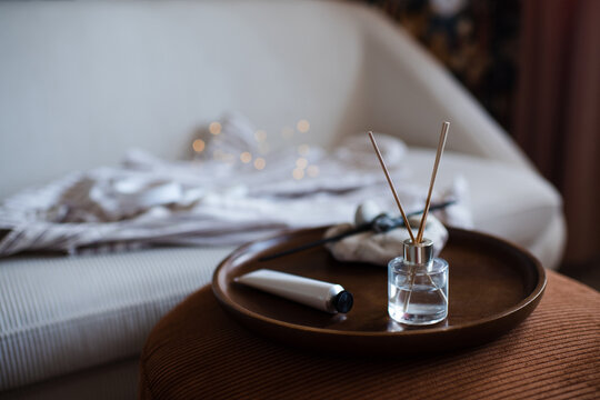 Home aroma liquid glass bottle diffuser with sticks on wooden tray and hand lotion in living room over couch. Hygge cozy atmosphere.
