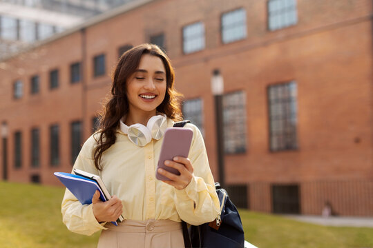 Smiling lady student with headphones around her neck, looking at smartphone while holding notebooks, enjoy chat, in sunlit college campus park