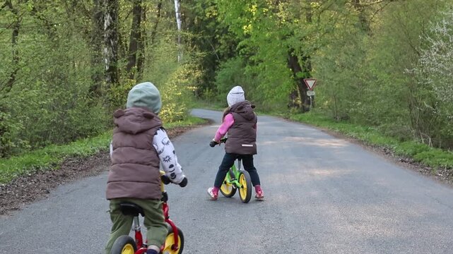 Two young children riding balance bikes down a forest road surrounded by colorful autumn trees.