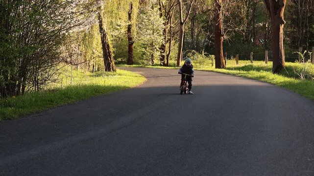 Adorable young child riding a red balance bike on a paved path through a sunlit park at sunset.