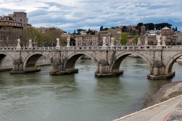Fototapeta premium View on stone-arched bridge on Tiber river in Rome, balustrade, marble statues, cloudy sky, ochre and terracotta buildings, domes and umbrella pines rise on gently sloping Vatican hill,