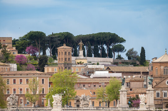 View on stone-arched bridge on Tiber river in Rome, balustrade, marble statues, cloudy sky, ochre and terracotta buildings, domes and umbrella pines rise on gently sloping Vatican hill, 