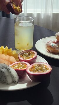 Tropical freshness and vitamins: woman scoops passion fruit pulp, mixes drink with spoon. Mango, papaya, jackfruit and donuts on buffet table. 
