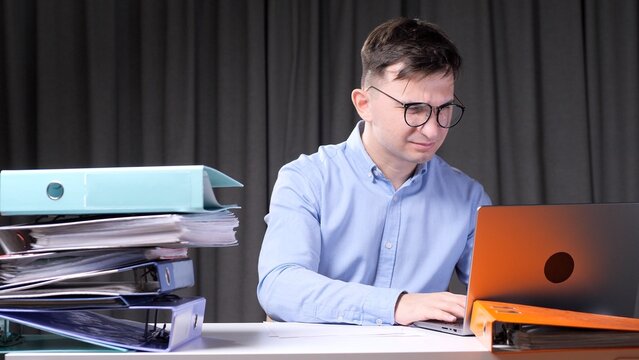 Stressed young office worker feeling frustrated while working on a laptop, surrounded by a tall stack of binders and documents, symbolizing workload and pressure