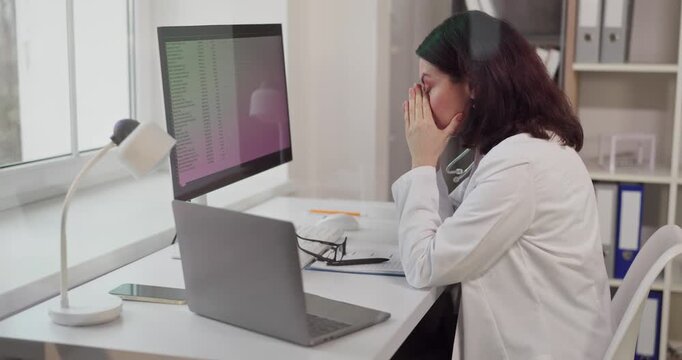 Doctor woman tired with headache at computer. In an office at a clinic she pauses by a monitor and laptop, managing records and paperwork under stress at work. Concept: medical burnout at work.