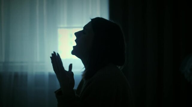 Young woman sitting alone in a dimly lit room, slowly raising hands in prayer near a window. Emotional scene of inner struggle, anxiety, loneliness and search for support, cinematic lighting and calm