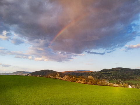 Aerial view of a vibrant green field and rolling hills under a cloudy sky with a rainbow in Cerin, Banska Bystrica Region, Slovakia.