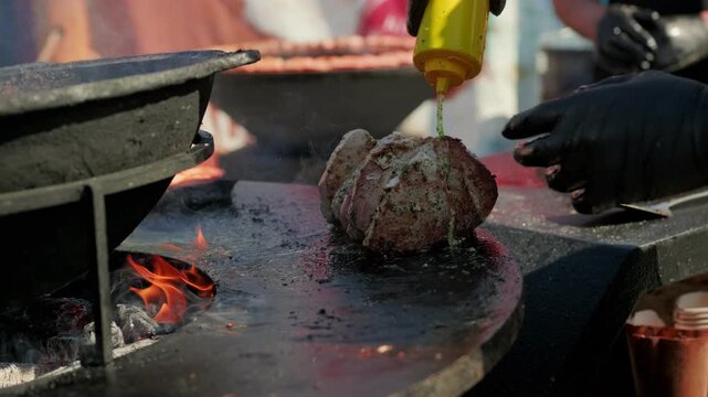 Chef grilling large piece of pork meat on open fire barbecue. Man cooking seasoned steak with metal utensil outdoors. Concept of street food and culinary skill.