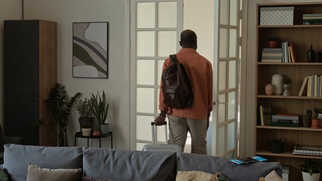Full shot of forgetful young African American couple with suitcases walking out of doors, departing for vacation, leaving behind tickets and passports on couch, man returning and picking them up