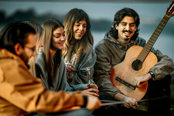 Naklejka premium Group of friends enjoying time together near water while playing music and having drinks during sunset
