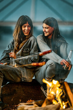 Two women enjoy roasting food over a campfire while sitting on a rock outdoors in the evening, sharing drinks and laughter together