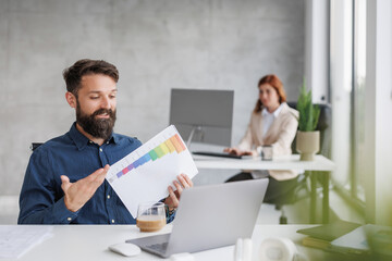 Fototapeta premium Man presents chart during meeting while woman works on laptop in modern office setting