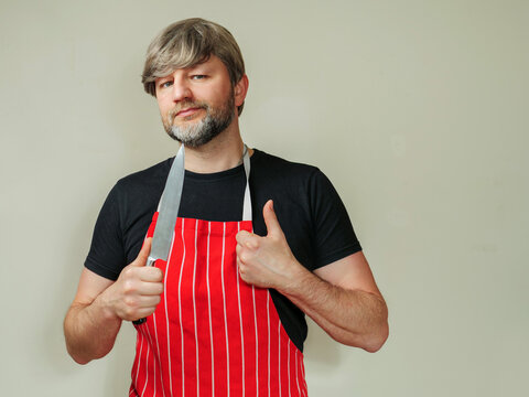Butcher of fish monger in black shirt and black and white classic apron on light background. Male in his 40s or 50s with grey hair and short beard holding knife. Mature professional.