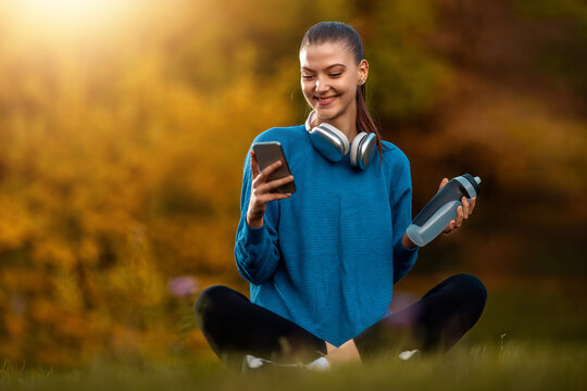 Young woman sits in park while looking at smartphone and holding water bottle during warm afternoon in autumn
