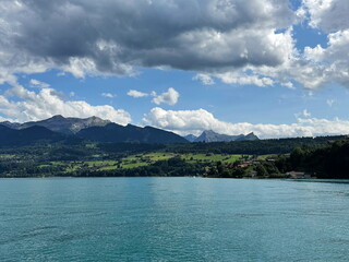 Obraz premium Blue waters of Lake Thun shimmer under bright clouds, with forested hills and distant mountains creating a peaceful summer landscape in Switzerland.