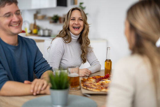 Friends gather for food and drinks while laughing at a cozy table in a home kitchen on a weekend evening