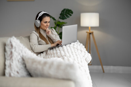 Woman uses laptop while sitting on sofa with blanket and headphones in modern room during daytime