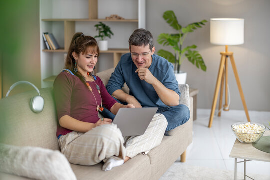 Couple sits together on a couch, working on laptop in a cozy living room with plants and snacks