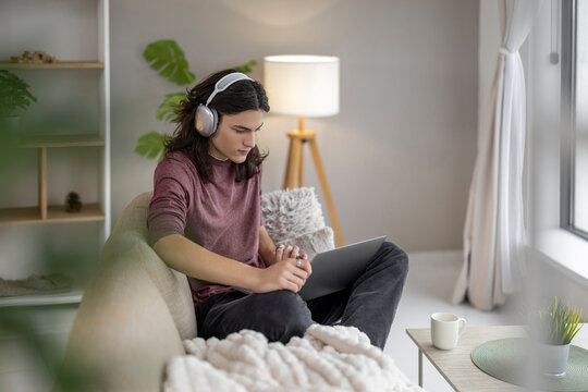 Young person sits on sofa using laptop in cozy living room while wearing headphones during day
