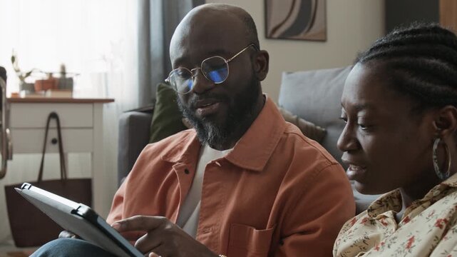 Medium close-up of African American couple sitting on floor by couch at home, researching resorts for luxury vacation on tablet, man describing hotel facilities while woman dangling credit card