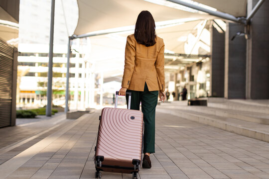 Young woman traveler walking away with suitcase in modern city environment