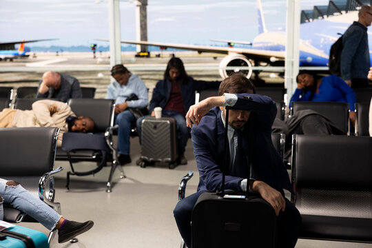 Tired CEO man in airport terminal lounge trying to rest during delayed flight business trip. Exhausted businessman sleeping on trolley handle, waiting for canceled flights to resume