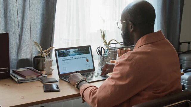 Medium rear shot of young black man sitting at desk, viewing exotic luxury resort on hotel booking website for summer holiday, scrolling down to check location map and taking out credit card to pay