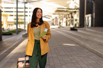 Obraz premium Young female smiling while holding passport and pulling suitcase in urban setting