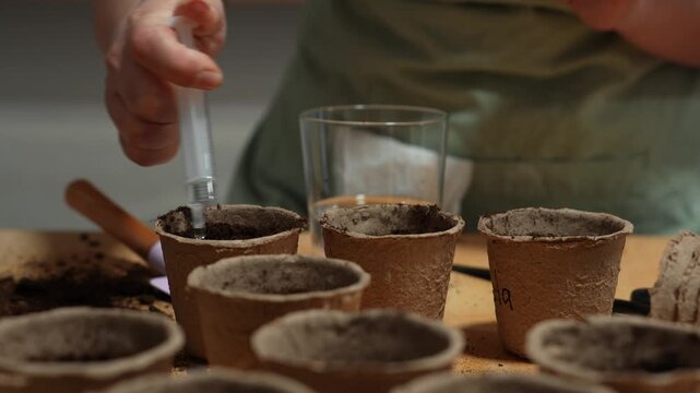 Gardener dispenses water droplets from a syringe into a prepared seedling container 4k video. Close-up of a gardener using a syringe to carefully water soil in a peat pot.