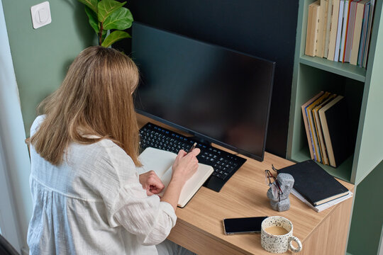 Woman writing in notebook at desk with monitor, keyboard, smartphone and books