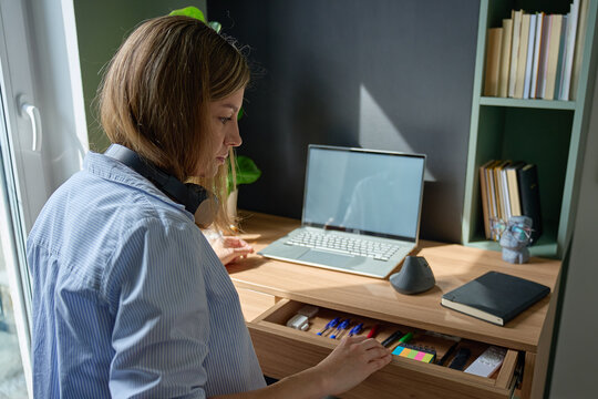 Woman sorting office supplies in desk drawer at home workspace with laptop