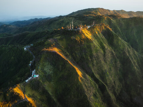 Aerial view of the lush mountain ridges of Bromo Tengger Semeru National Park with golden sunlight hitting the slopes and communication towers on the peak in Bromo, East Java, Indonesia.