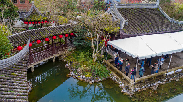 Aerial view of Lan Su Chinese Garden featuring traditional tiled roofs, red lanterns over a bridge, a tranquil pond, and visitors gathered under a white tent in Portland, Oregon, United States.