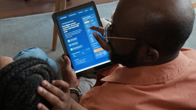 High-angle over-shoulder shot of young African American couple choosing vacation flights online on tablet using travel booking aggregator website, discussing dates, prices and options