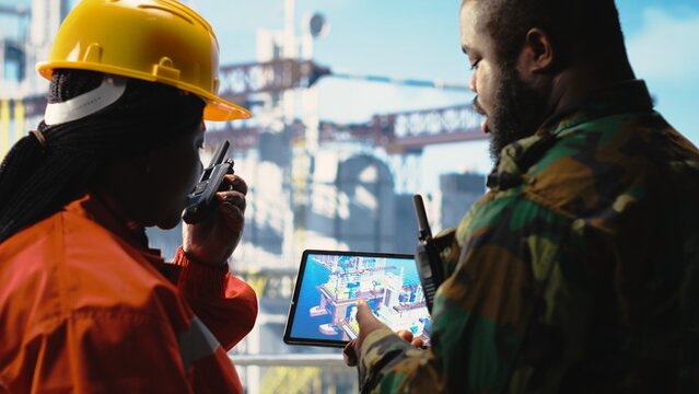 Soldier does escort duty for offshore platform vessels, verifying system integrity using device app. Army unit guards drilling rig against terrorist threats using tablet next to engineer