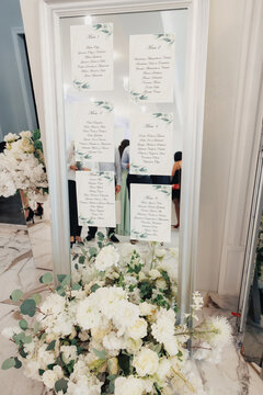 Mirror Seating Chart With White Florals, Handlettered Calligraphy On Glass Panels, Gold Frame Anchored Among Lush Roses And Peonies, Marble Floor Reflection, Scattered Petals, Soft Natural