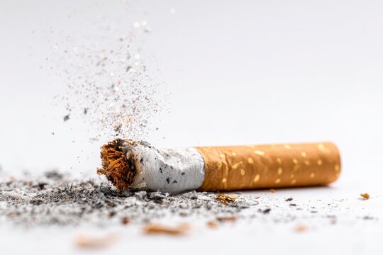 Lit cigarette butt lying on a white surface, sparks and ash scattering around, macro shot of tobacco combustion.