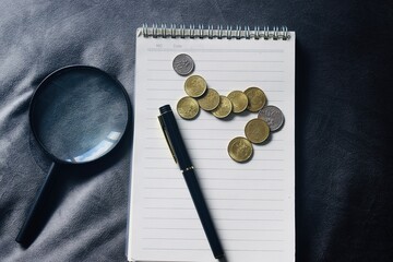 Top view of a magnifying glass, a black fountain pen, and scattered Malaysian Sen coins on a white spiral notebook. © Akram