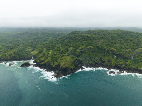 Aerial view of Bukit Pengilon featuring lush green hills, rocky cliffs, and a small waterfall cascading into the turquoise ocean in Kecamatan Tepus, Daerah Istimewa Yogya, Indonesia.