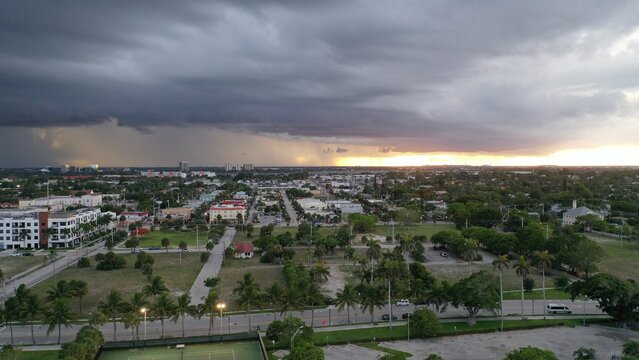 Aerial view of a suburban neighborhood under a dramatic storm front with dark clouds, falling rain, and golden sunset light on the horizon.