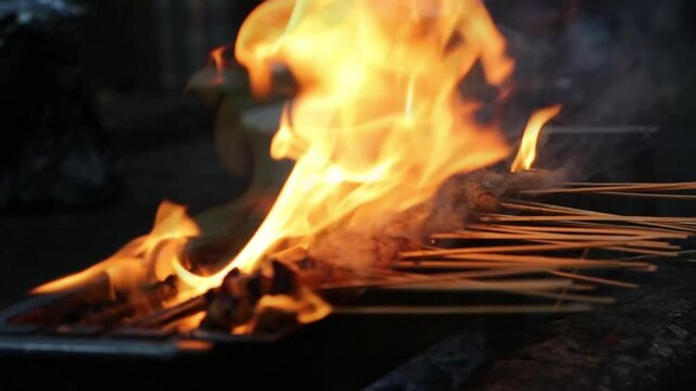 Flaming satay skewers grilling over open fire, smoky Asian street food BBQ with dramatic orange flames and cinematic night cooking.
