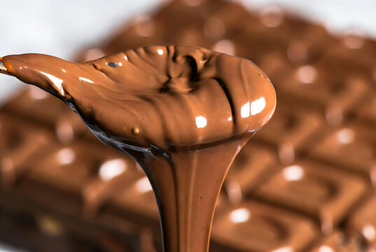 Melted chocolate is being scooped from a spoon with a chocolate bar in the background. The chocolate is smooth and rich.