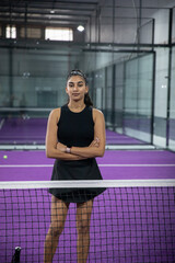 Fototapeta premium Indian woman standing behind net on purple glass court, wearing black skort, watch, ball on left
