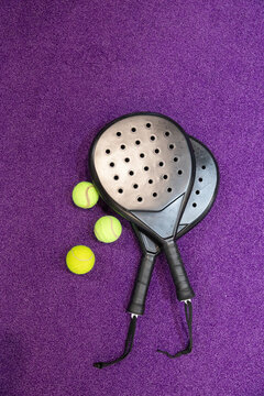 Two black holed padel paddles crossing purple court with black grips or straps, 3 yellow tennis ball