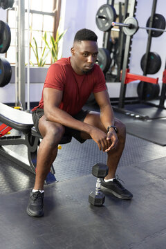 African American man sitting on bench in gym, resting between sets with dumbbell, wearing red shirt